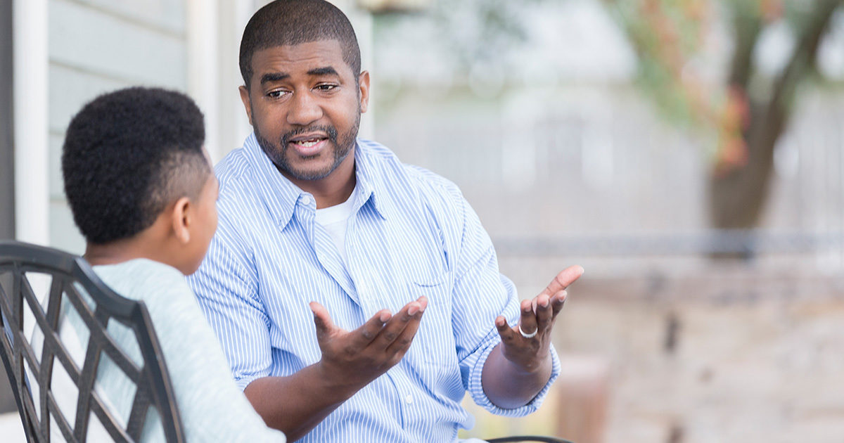 A middle-aged African-American man sits speaking to his son. The man's hands are held open in an expressive gesture.