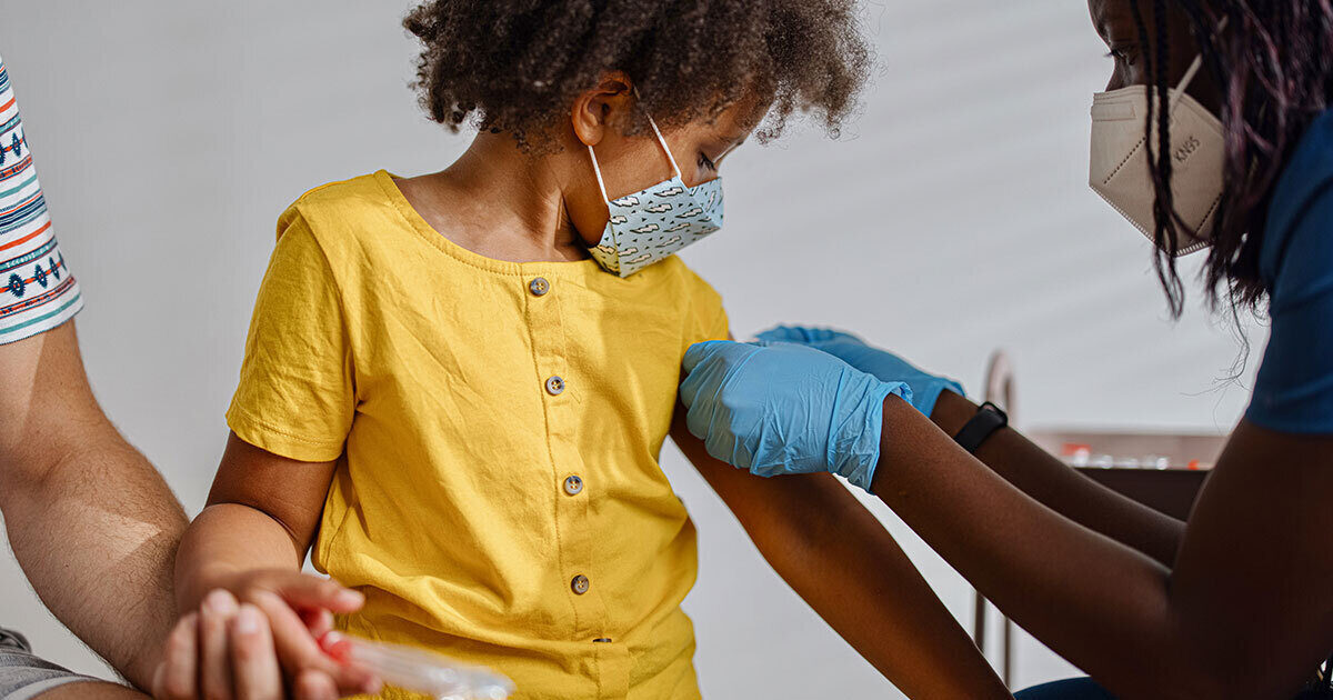 A young girl wearing a yellow button up shirt holds her parent's hand as a provider wearing blue latex gloves puts a bandaid on her arm where she just received a vaccination.