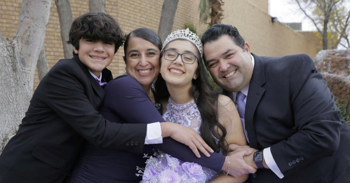 The Candelaria family is standing outside, hugging, and smiling for the camera at Patty's quinceañera.  Patty is wearing a lavender colored dress with a tiara.