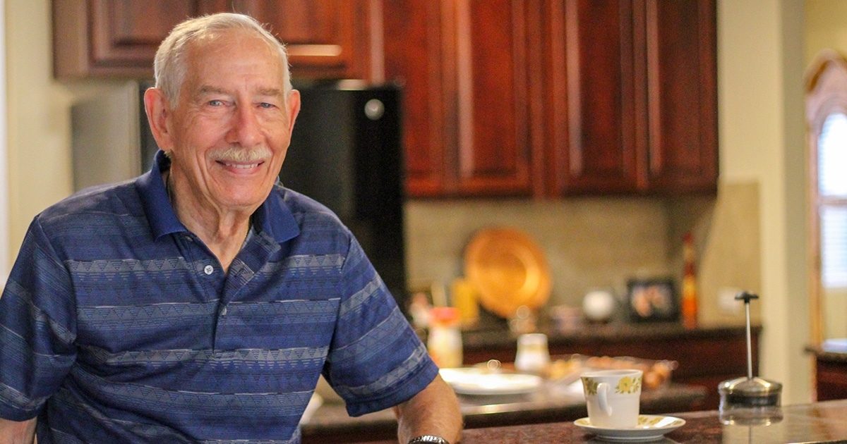 A senior man with a mustache stands in a kitchen, his arm resting on a countertop. He is smiling at the camera and wears a blue striped polo shirt.