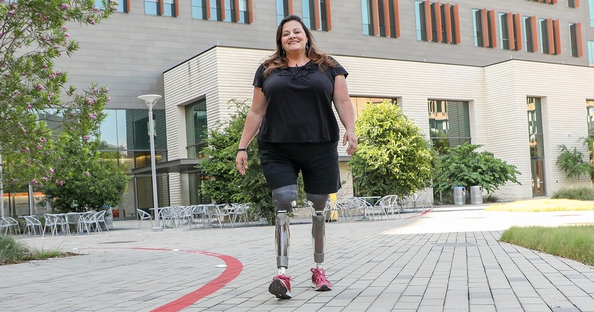 Heather, a patient with UT Health Austin's Musculoskeletal Institute, is seen walking with prosthetic legs across the courtyard at UT's Health Transformation Building.