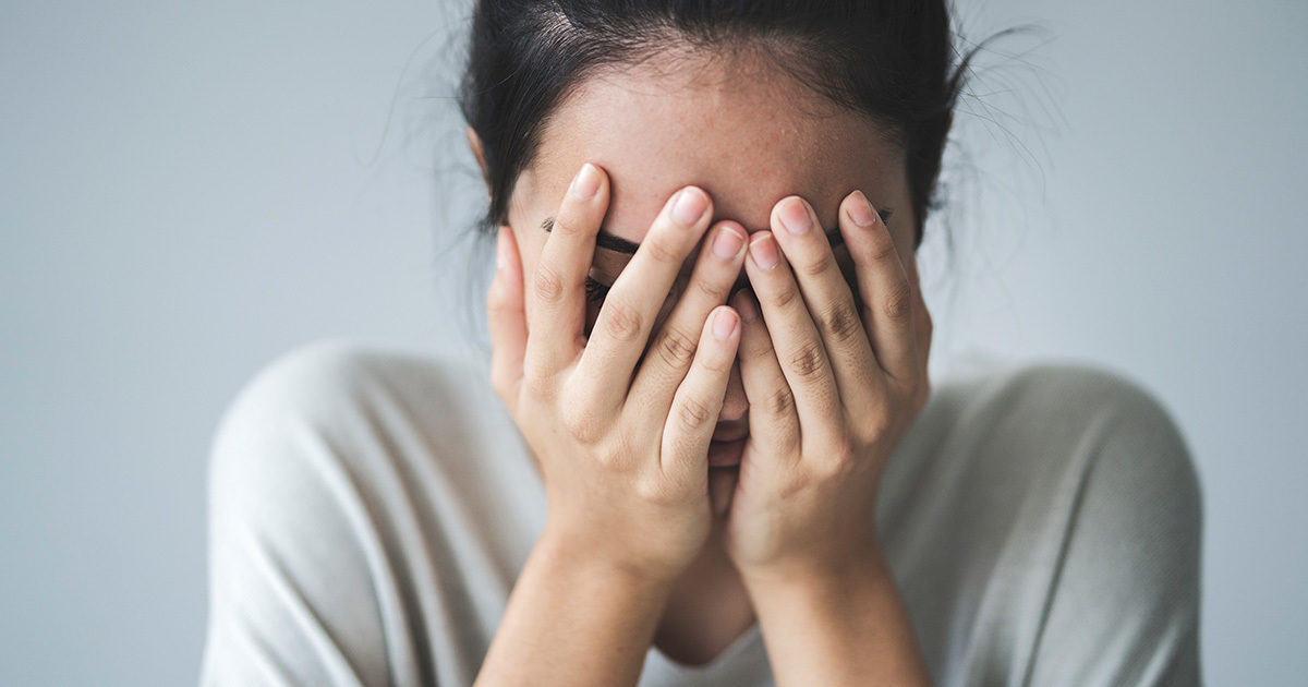 A young woman faces the camera with her face buried in her hands.