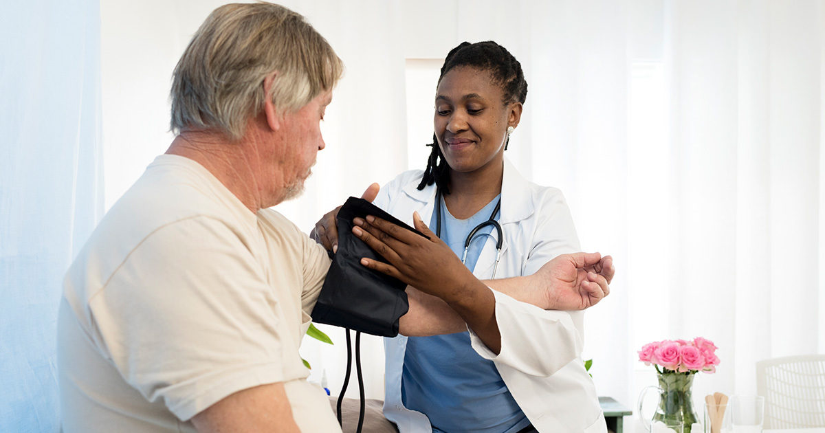 A female, African-American clinician places a blood pressure cuff around the arm of an older, White man.