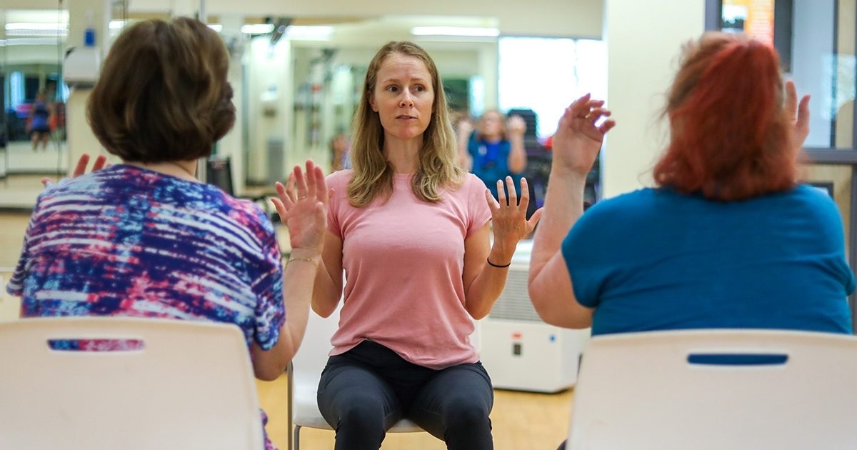 Two women sit in chairs, backs facing the camera, raising their arms in an exercise. Between them sits another woman, facing the camera, guiding them through the movements.