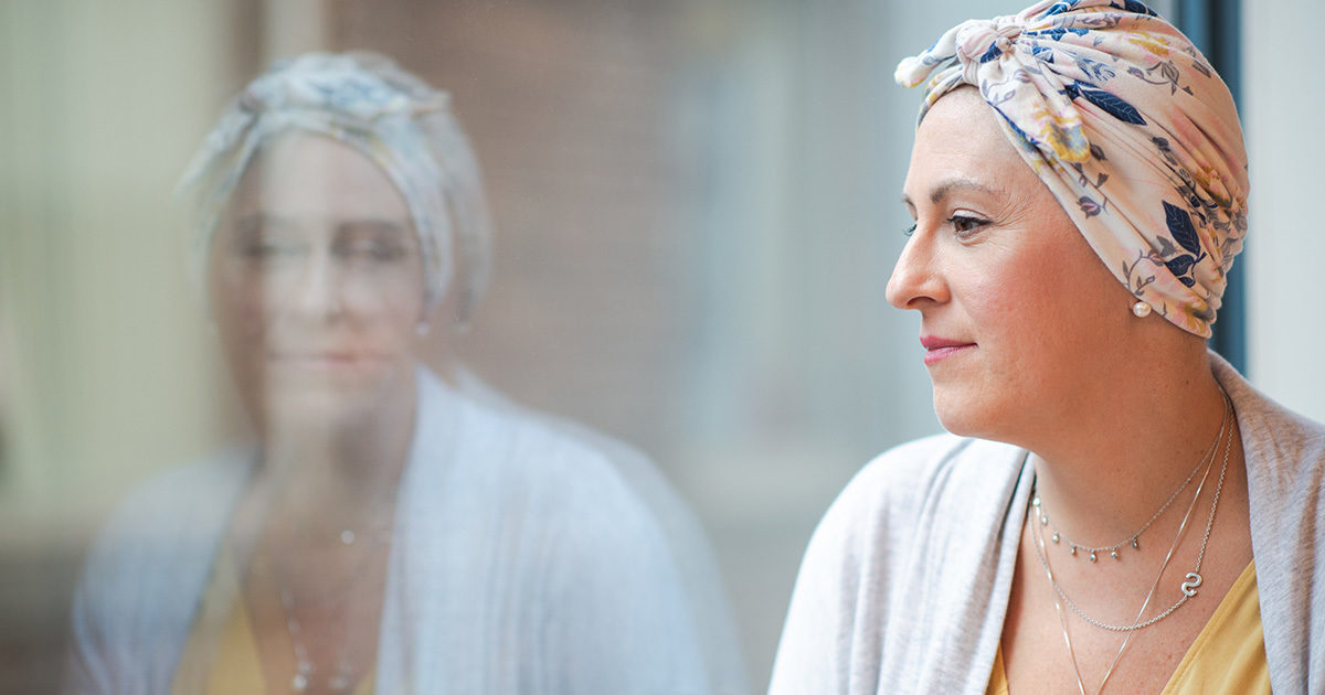 A woman wearing a head scarf stares out of a window. Her image is reflected in the glass.