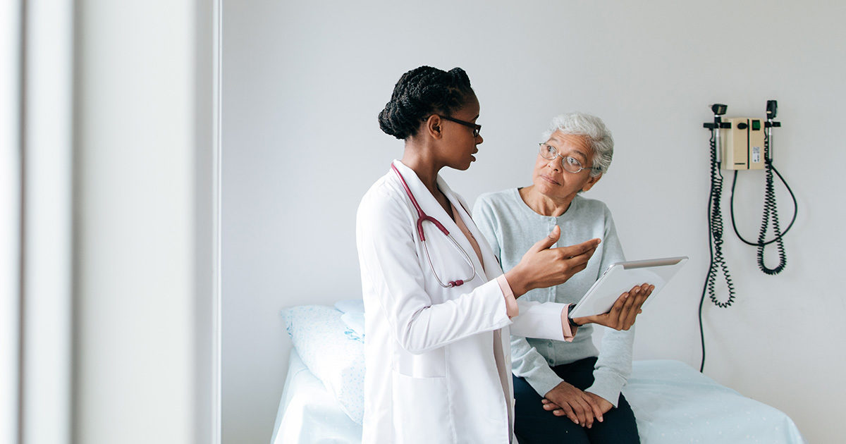 A senior African-American woman sits on an exam table listening as a young Africa-American clinician in a white lab coat speaks with her.