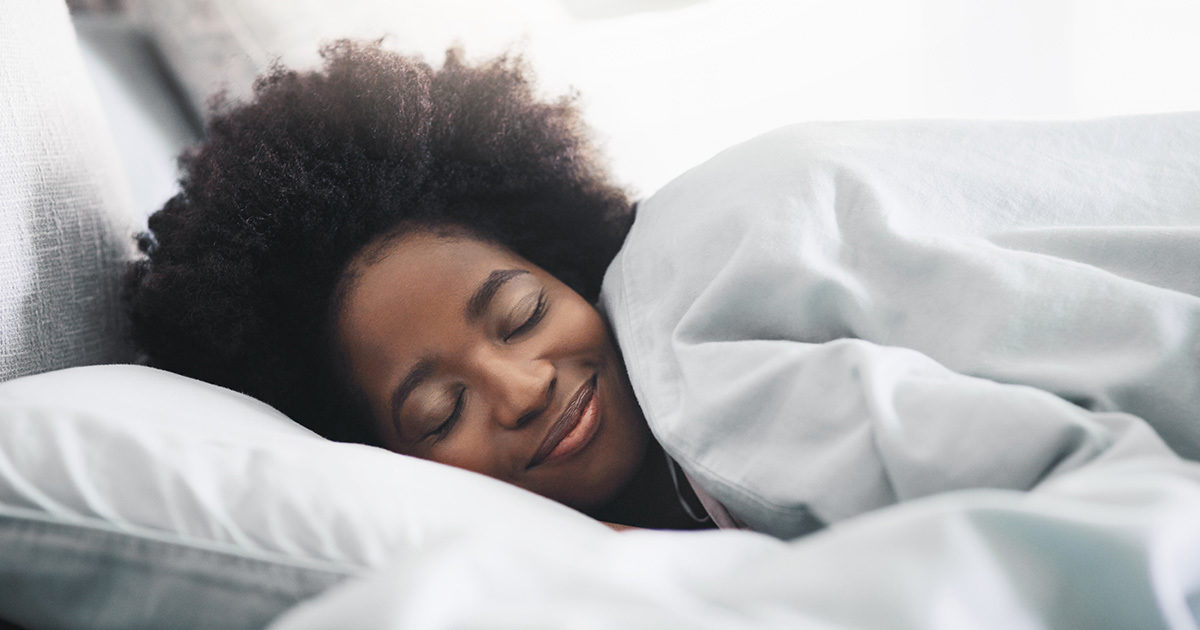 A young African-American woman is shown asleep with her head on a large white pillow. A pleasant expression is on her face.