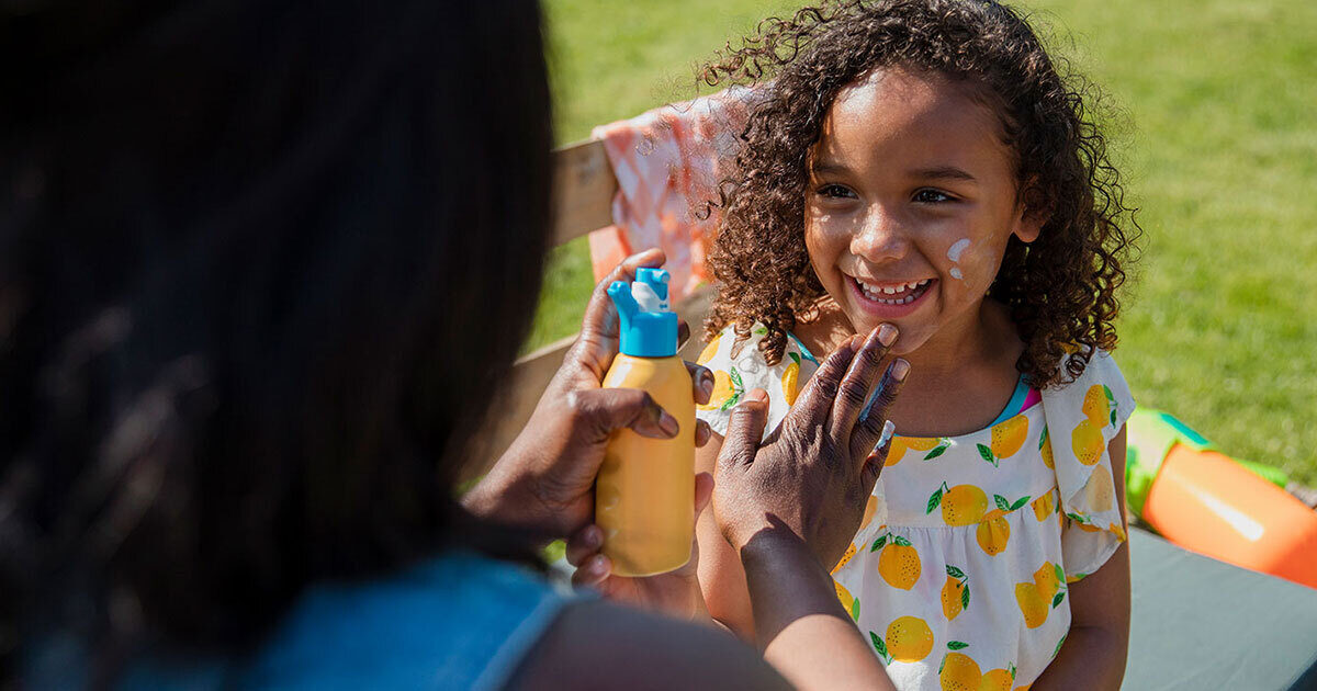 A woman applies sunscreen to her daughter's face as they sit outdoors on the grass.