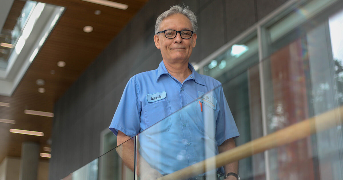 Eduardo Garea, the Crew Leader of Facilities Services, smiles down from a glass balcony. He is wearing a custodial uniform and glasses.