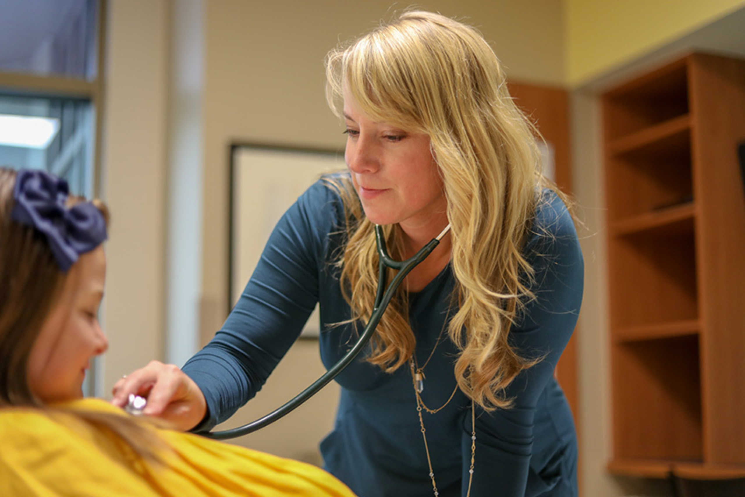 Photo of a young girl, dressed in a yellow shirt and navy blue hair lays down on an exam room while a clinician listens to her heart with a stethoscope.