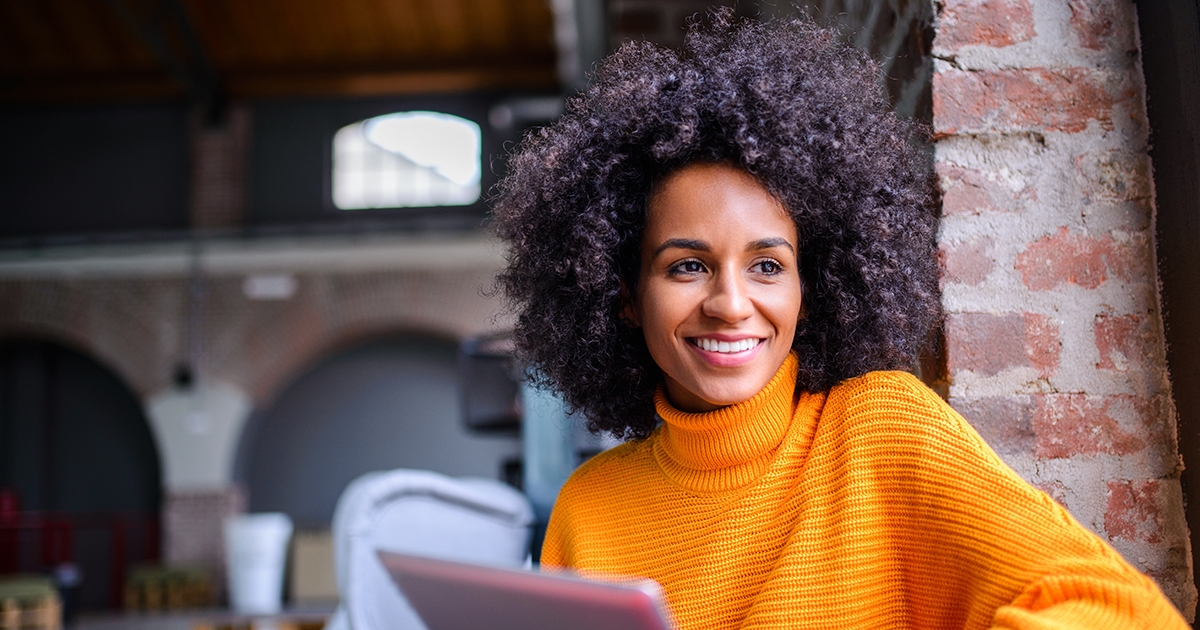 A young African-American woman sits at a table with a laptop, smiling at the camera.