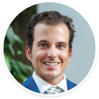 Urologic oncologist Aaron Lavinia, MD, MBA, wearing a suit and tie and smiling near a white wall and a tree.