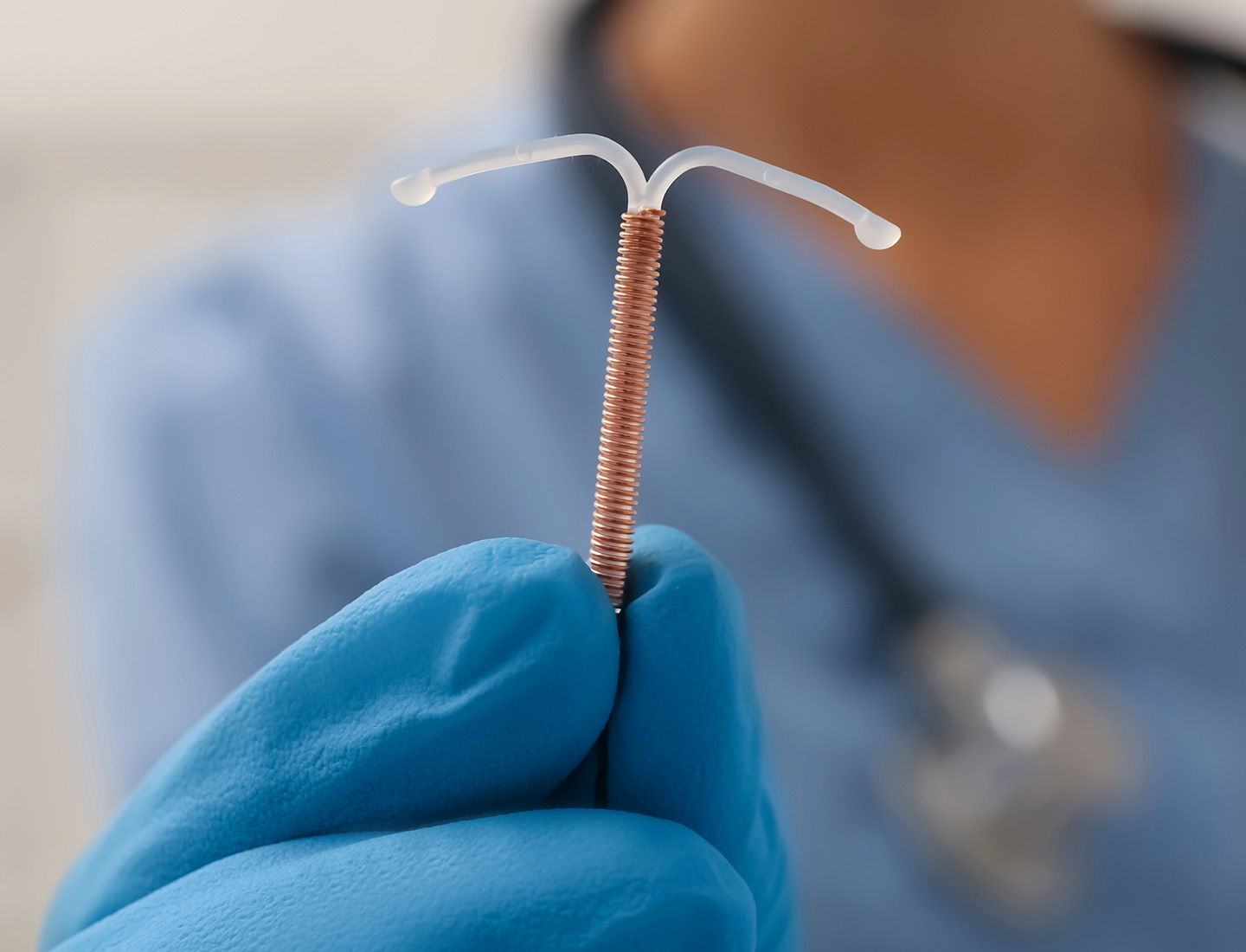 An IUD device is close up and in focus while a person wearing a stethoscope, surgical gloves, and blue scrubs is blurry in the background.