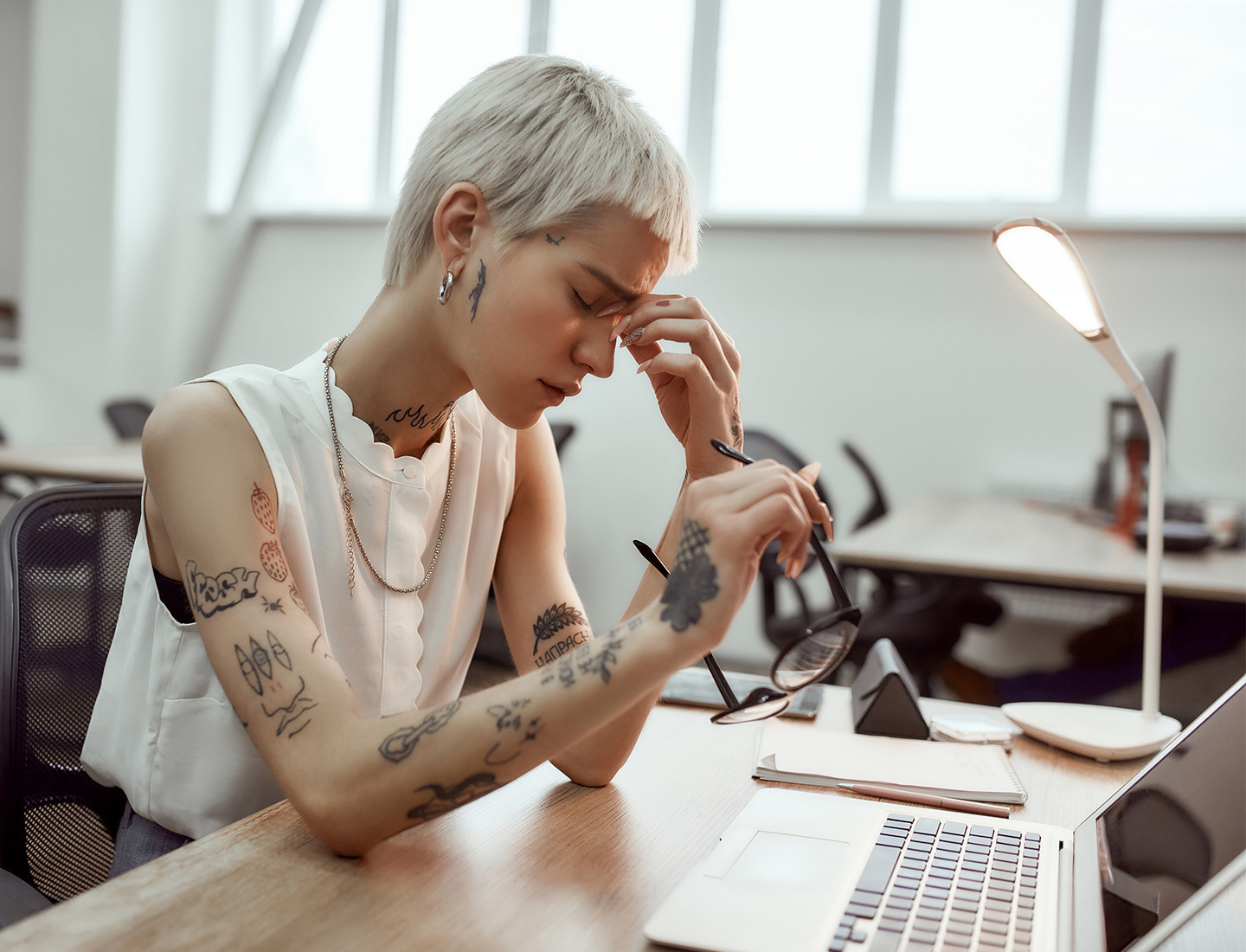 A woman sitting at a desk with a laptop squints and pinches the bridge of her nose, holding her glasses with her other hand.