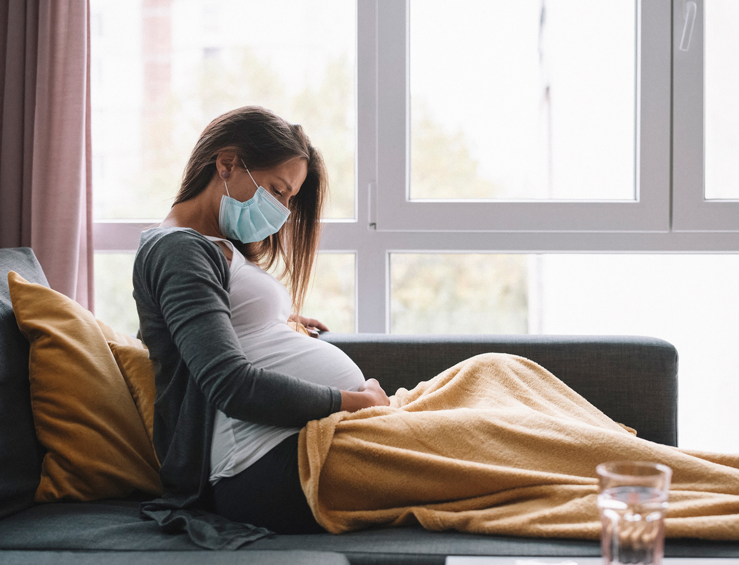 A young adult pregnant women, wearing a face mask, sits on her couch covered in a blanket.
