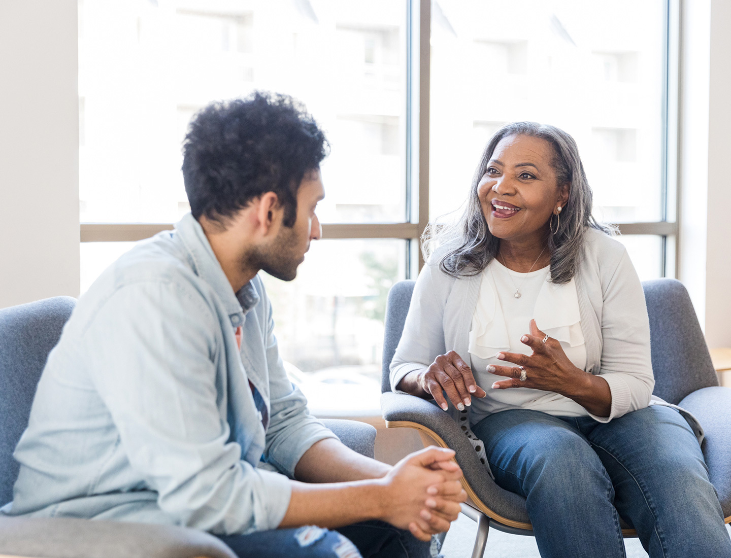 A young man and a woman with gray hair seated in blue chairs engaging in conversation.