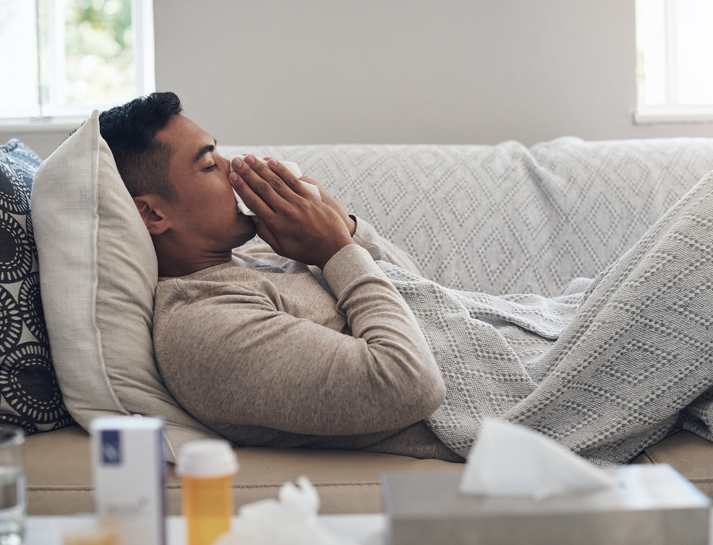 A young man lying on a couch, holding a tissue to his nose and mouth. He is covered in a throw blanket and medicine and tissues are visible on a coffee table in the foreground.