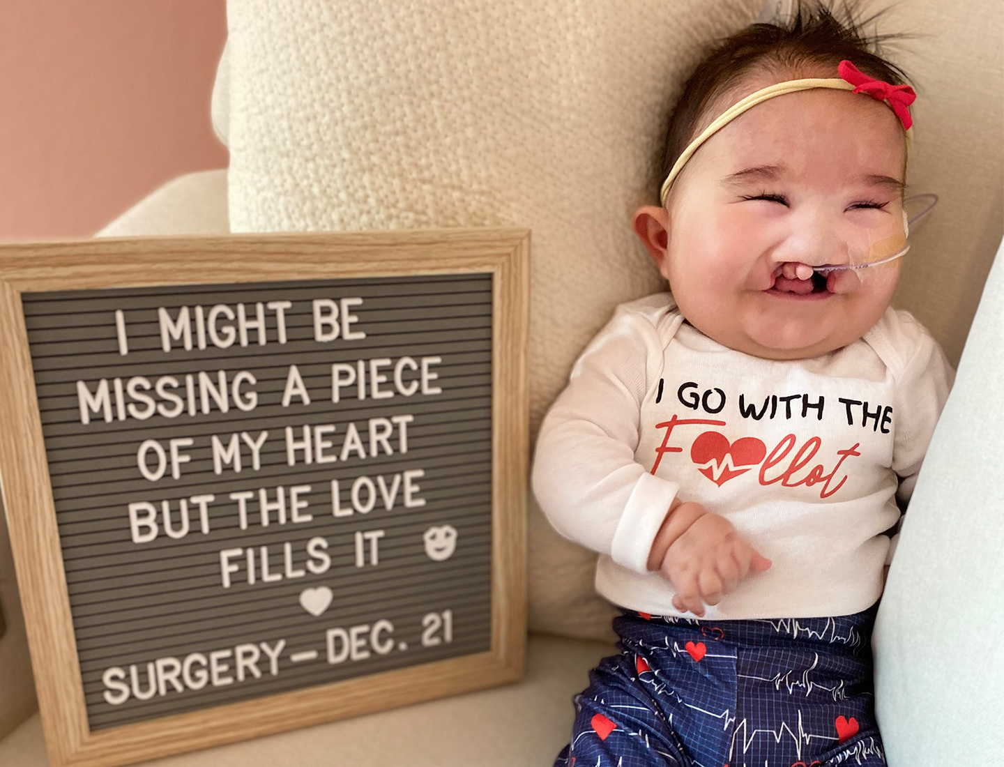 Baby Ruby Sedillo posed next to a gray letter board that reads "I might be missing a piece of my heart but the love fills it."  Her shirt says "I go with the Fallot", her pants are printed with hearts, and a red bow is attached to her headband