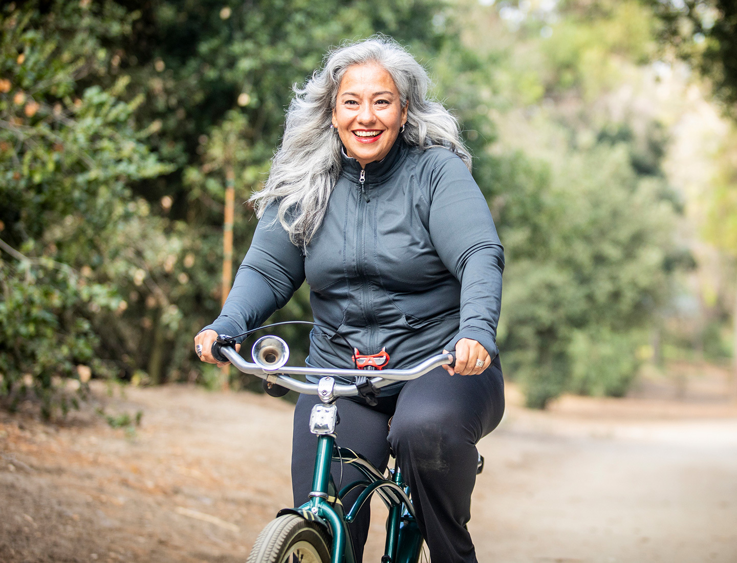 A smiling woman with gray hair rides a bike on a path lined with trees.