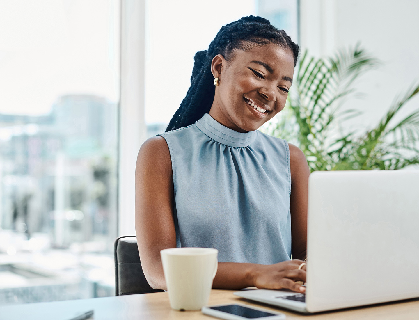 A young woman in a blue shirt tying at a laptop at her desk with a cup of coffee nearby. Her environment is brightly lit by a large window.