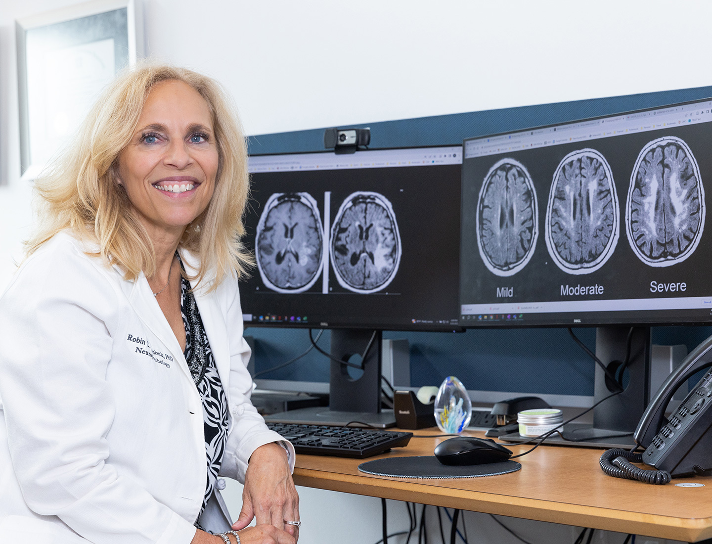 Robin Hilsabeck, PhD, ABPP, seated in front of two computer monitors displaying MRI brain scans.