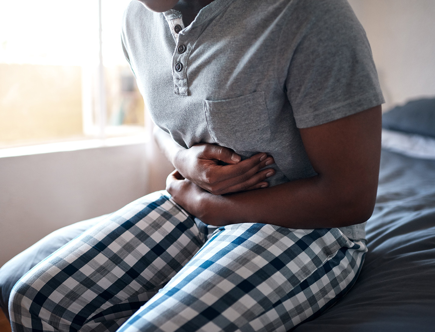 A man dressed in a gray shirt and blue and green flannel pants sits on the end of his bed, leans forward and holds his stomach with both hands.