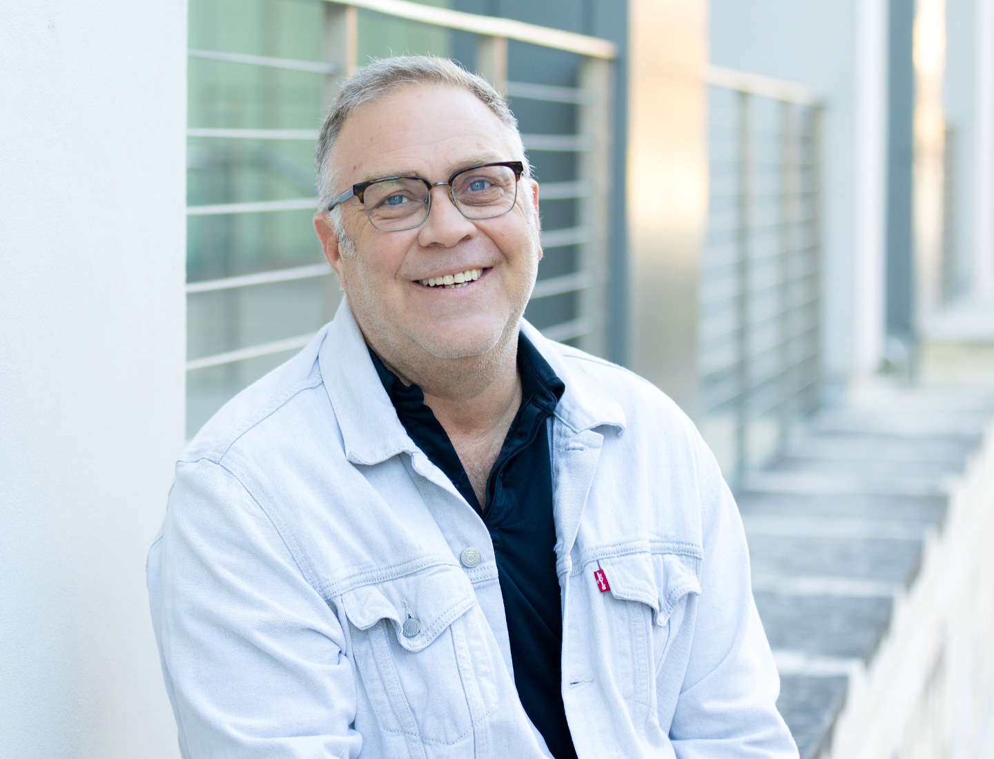 Jason Dudey smiling outside the Health Transformation Building at UT Health Austin. He is wearing a denim jacket and dark polo.