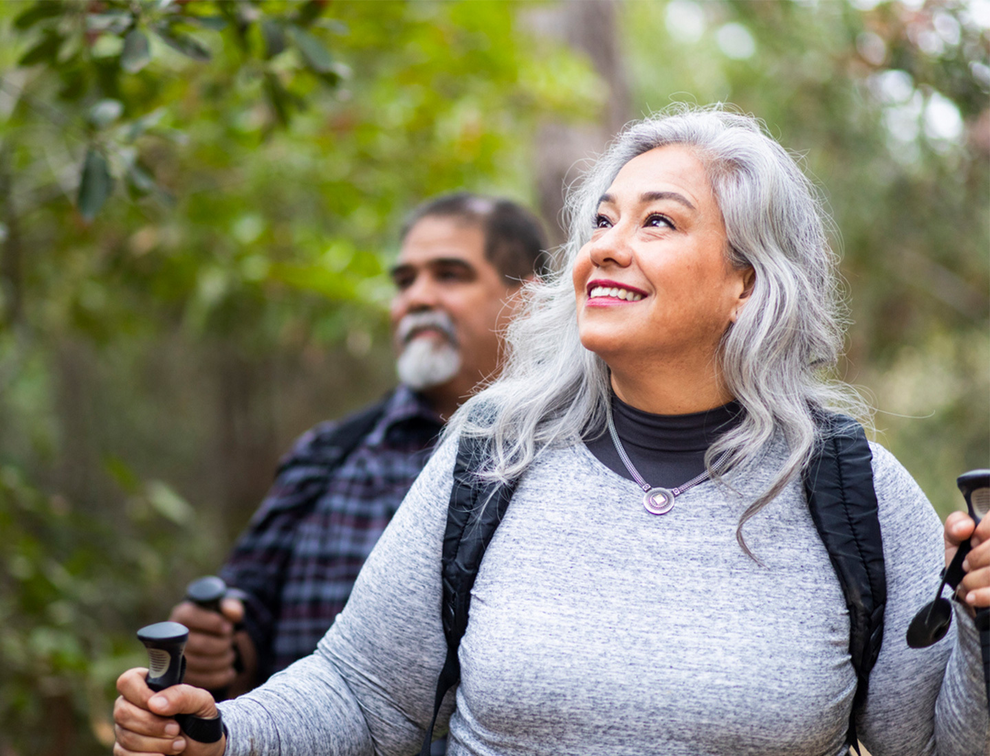 A woman with gray hair using walking sticks to hike through a forest. Behind her is a man with dark hair and a gray beard.