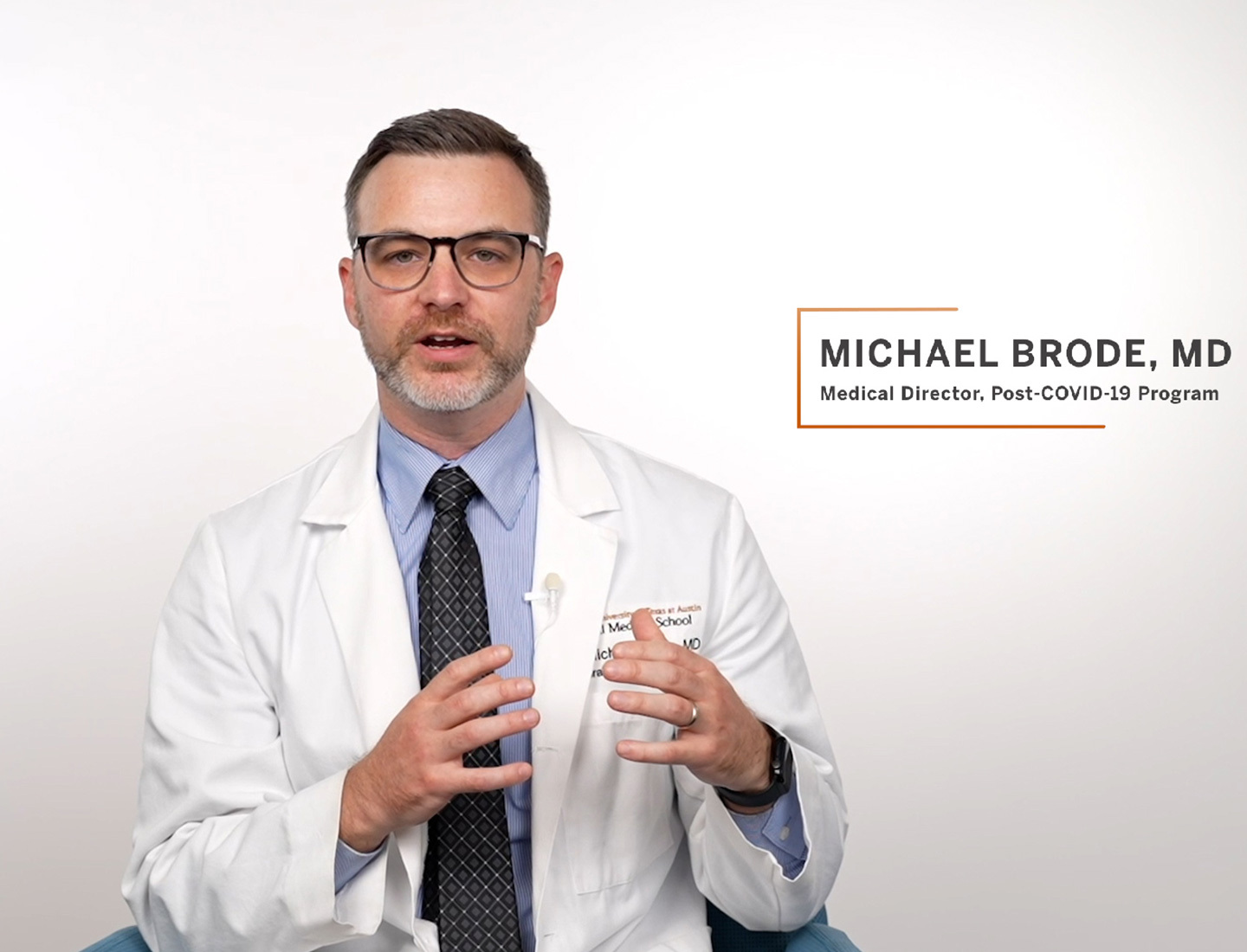 Michael Brode, MD, the Medical Director of the UT Health Austin Post-COVID-19 Program, wearing a white coat and speaking in front of a white backdrop.