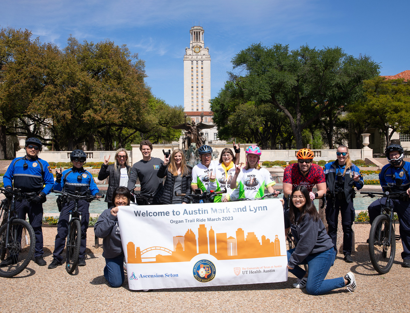 Participants in the Organ Trail ride through the UT Austin campus posing in front of Littlefield Fountain with a banner reading "Welcome to Austin, Mark and Lynn. Organ Trail March 2023." The UT tower is visible in the background.