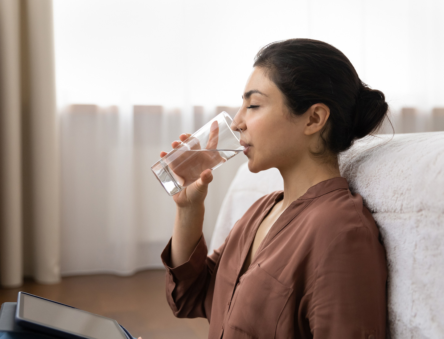 A woman sits on the floor of her bedroom, leans against her bed, and drinks from a glass of water.