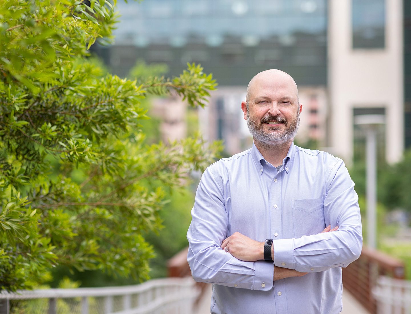 Dr. Jared Benge is standing outside of the HTB on a bridge surrounded by green foliage.  He is crossing his arms, smiling, and wearing a light purple shirt.