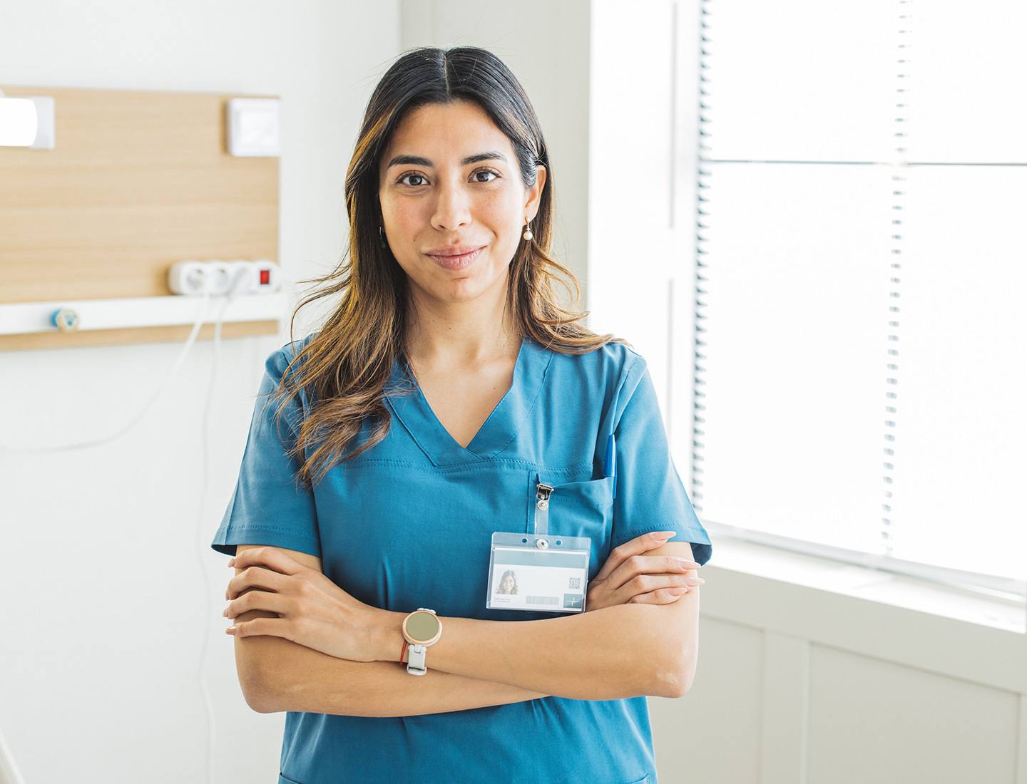 A long-haired woman with teal scrubs and a name badge crossing her arms and smiling near a window.