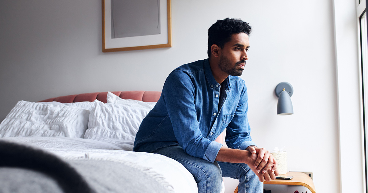 A young man dressed in a blue shirt and jeans, sits on the edge of a bed with hands crossed, looking out a window.