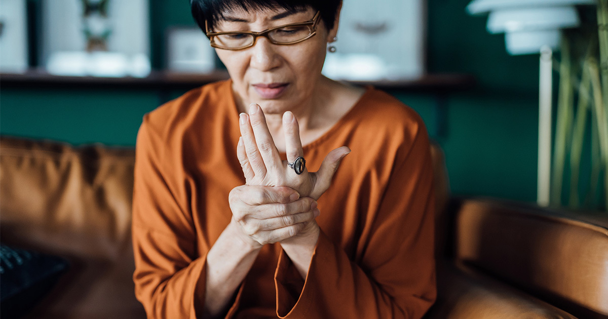 An older woman holds her wrist in her other hand while sitting on the couch.
