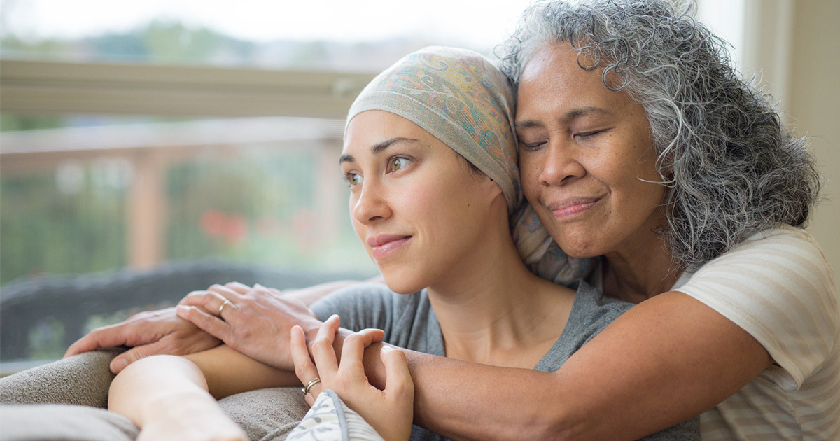 A young woman with cancer in a head scarf is being hugged around her shoulders from behind by an older woman with her eyes closed.