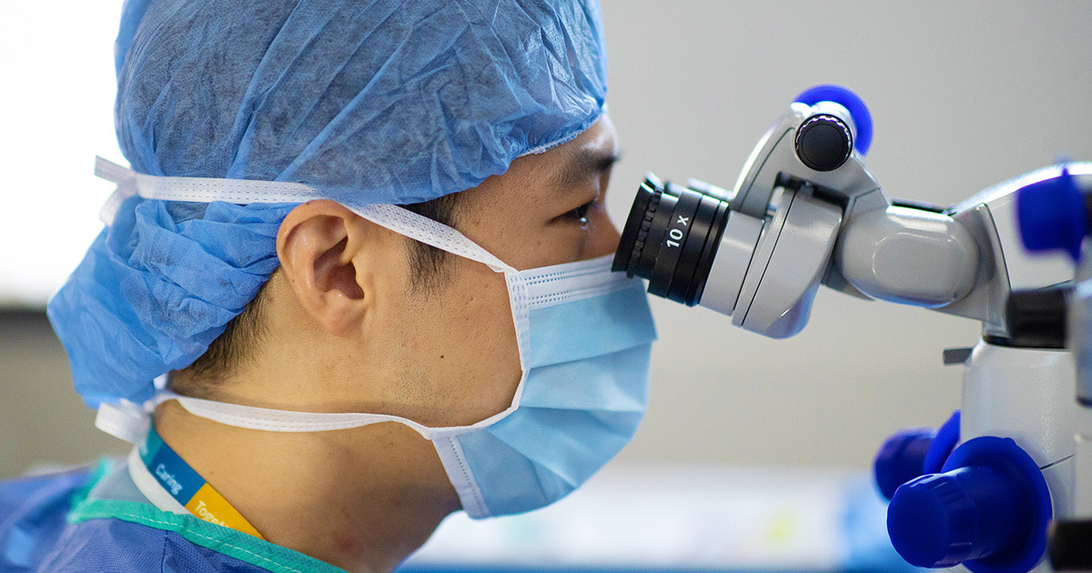 Ophthalmologist Gene Kim, MD, in a scrub cap and mask, peers into a microscope during a surgical procedure.