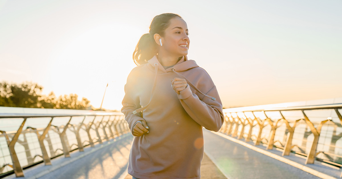 A woman wearing a brown sweatshirt and wireless earbuds jogging across a bridge.