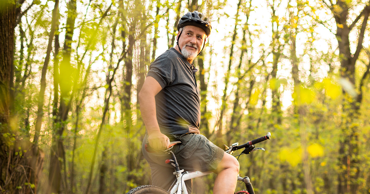 A middle-aged man riding a bike wearing a helmet is stopped in a forest and has turned back to look at the camera.