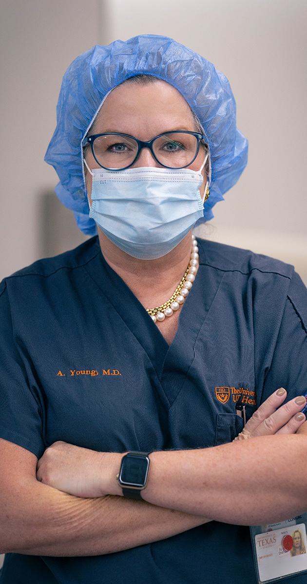 Amy Young, MD, posing in the operating room in dark scrubs. Her hair and her face are covered by personal protective equipment.