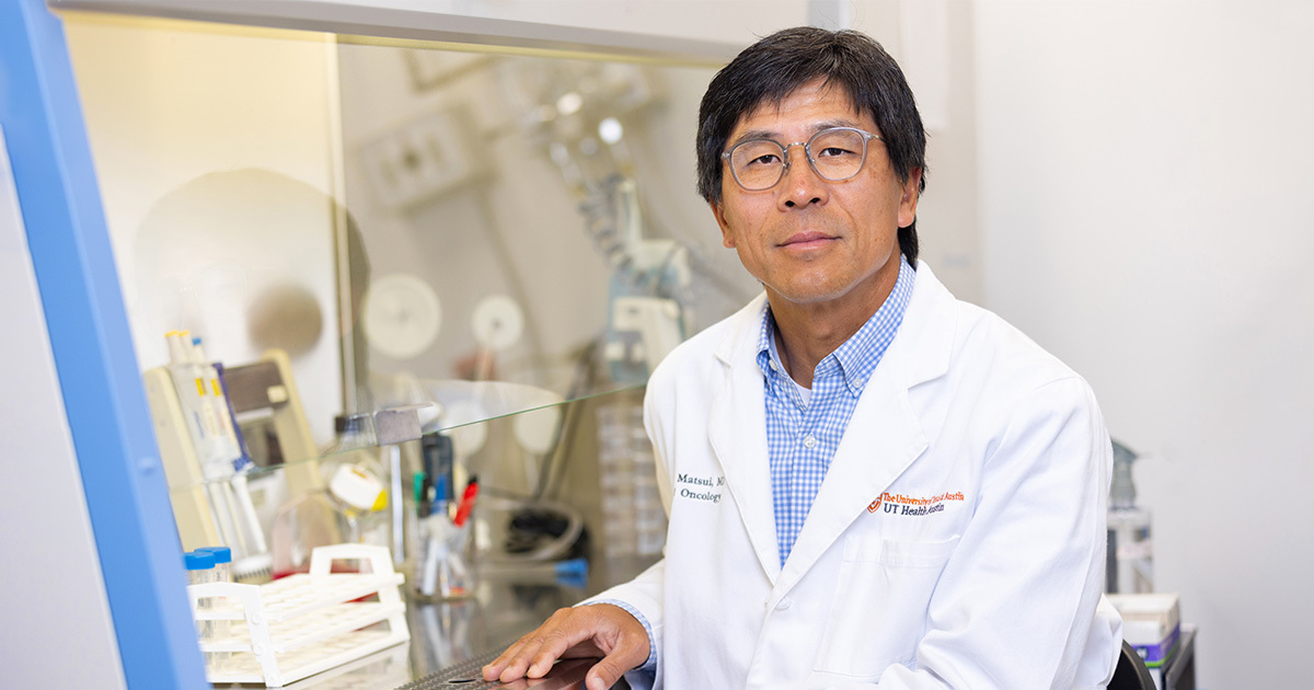 William Matsui, MD, posing in a white lab coat next to a fume hood full of laboratory equipment.