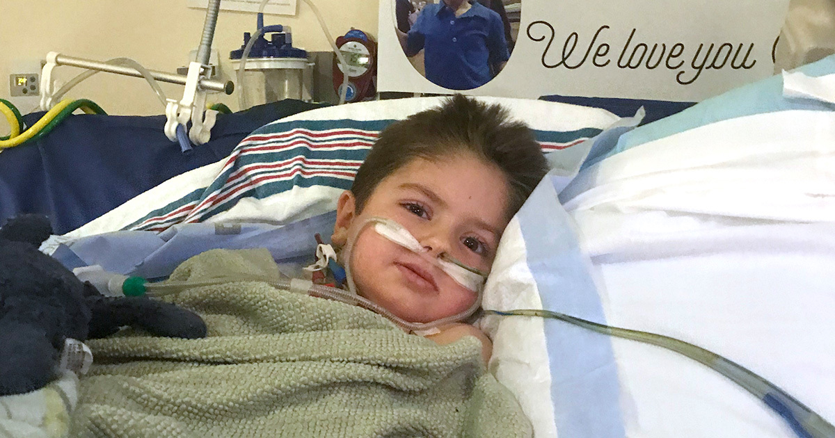 Owen Coulter lies in his hospital bed wrapped in an olive green blanket with a sign behind him that reads, "We love you".