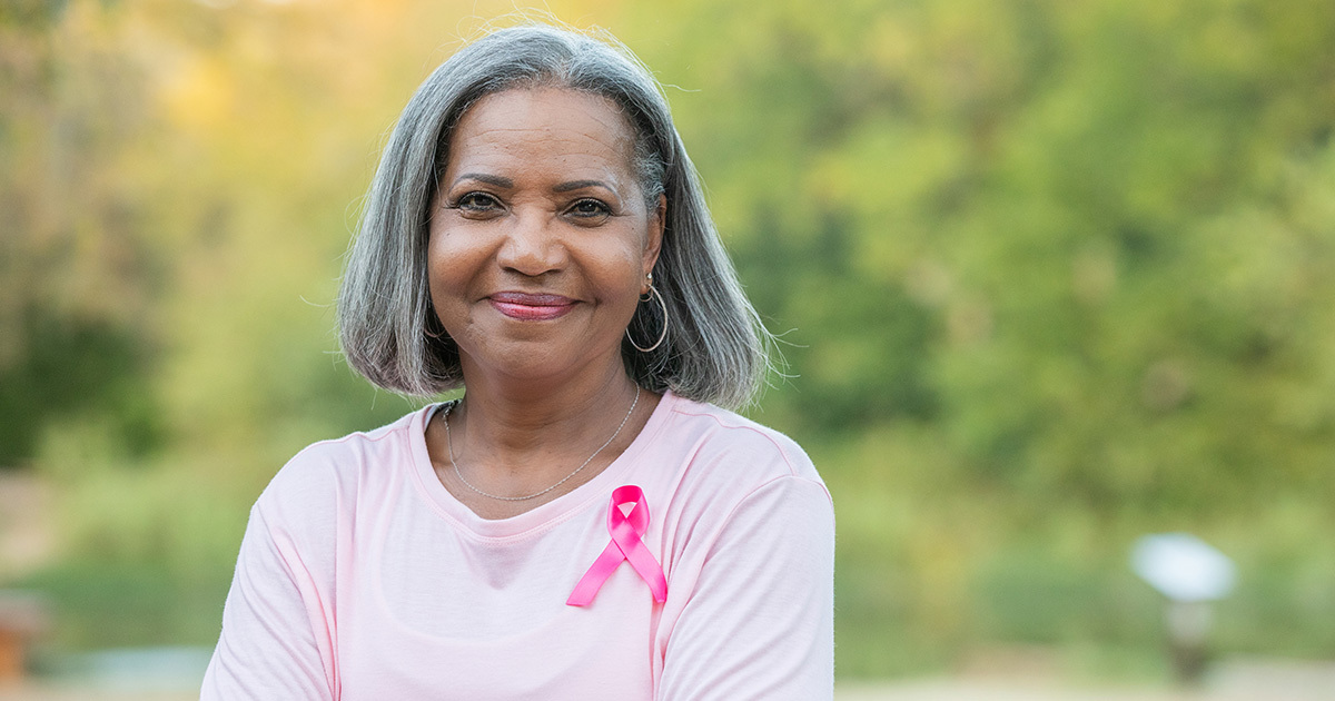 A woman with short gray hair and a pink breast cancer awareness ribbon pinned to her pink shirt standing outdoors