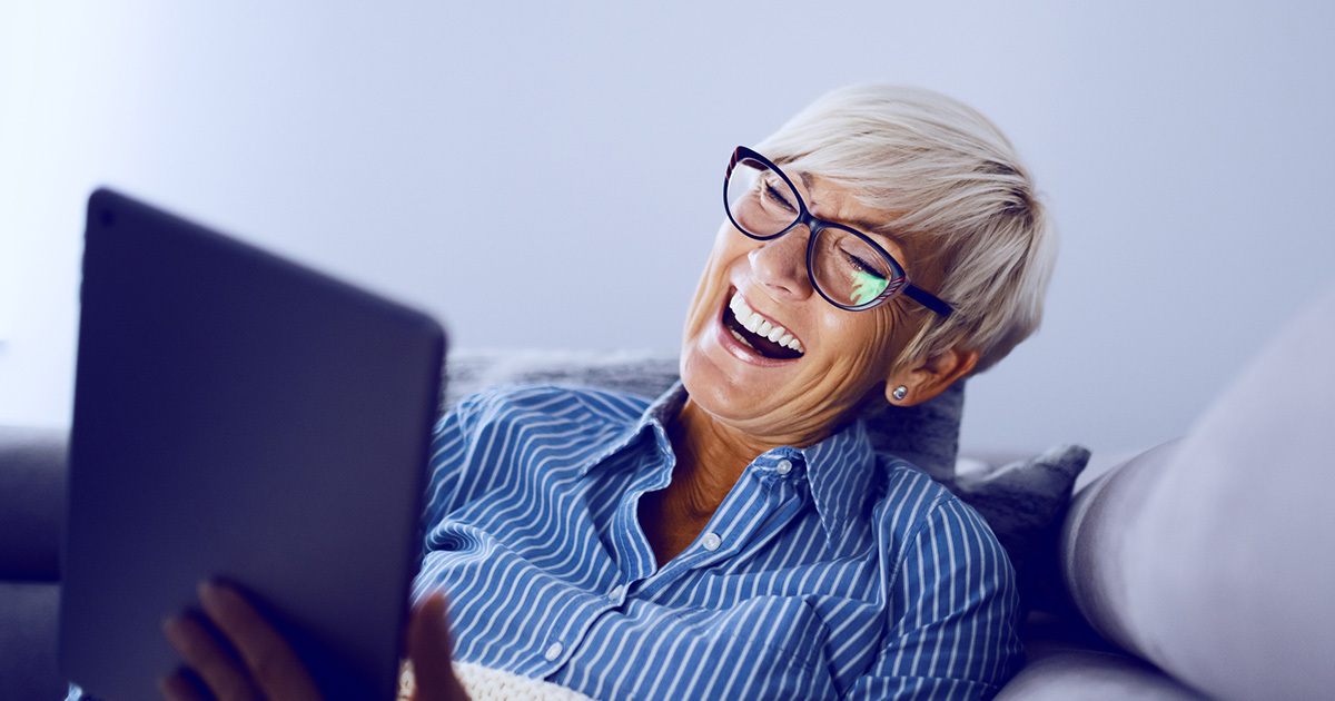 An elderly woman with short silver hair and blue glasses is sitting on a couch, smiling and laughing at her ipad.