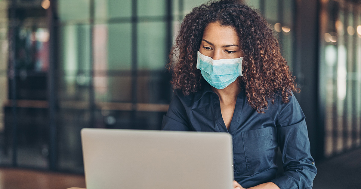 A young woman with curly hair and wearing a mask sits at a table working on a laptop computer.