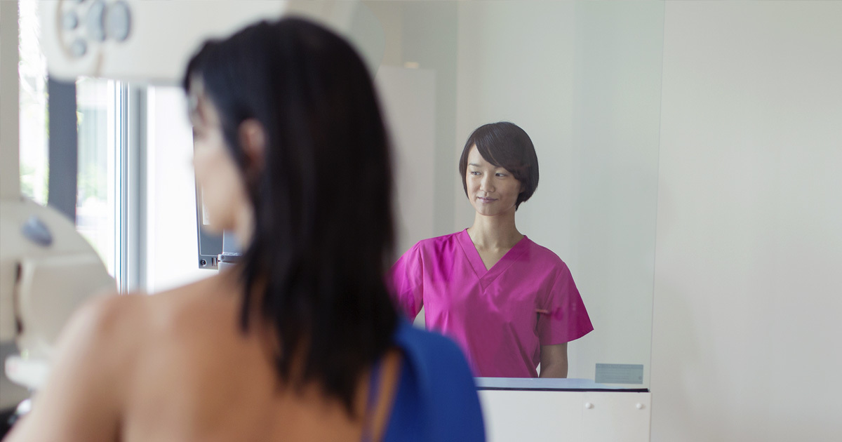 Out of focus in the foreground is a woman undressed for a mammogram. A nurse is visible in a nearby mirror.