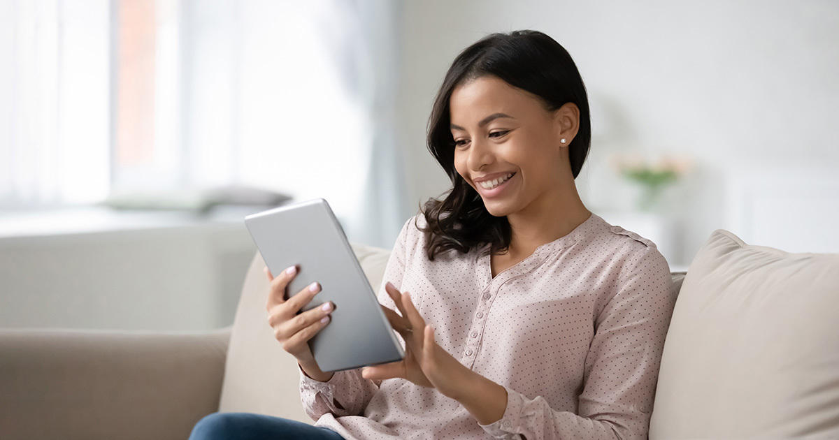 A woman sitting on a couch, holding a tablet while reading and smiling.