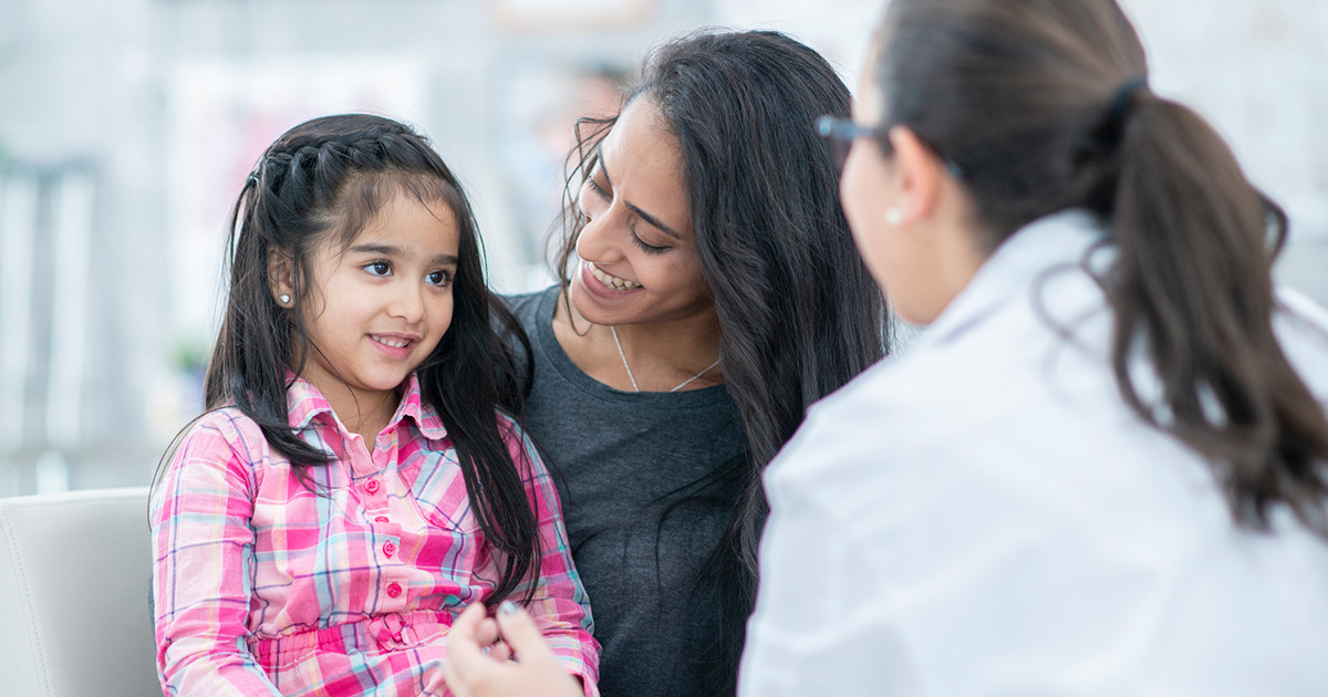 A young pre-adolescent girl sits next to her mother while engaging with a medical provider.