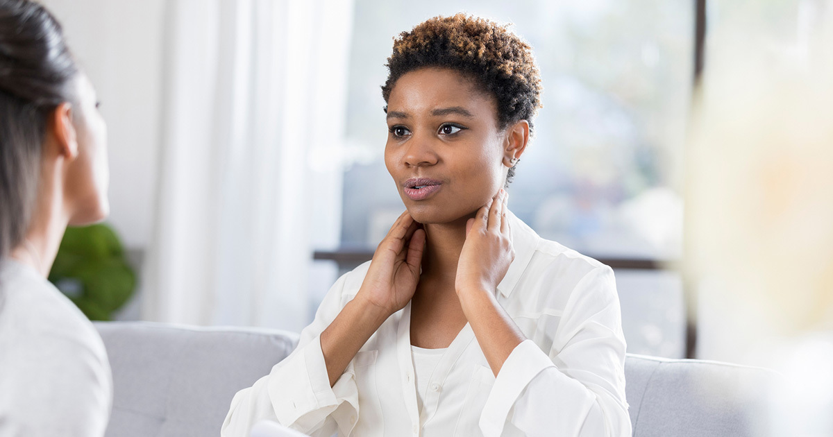 A young woman is shown with her hands on either side of her esophagus talking to a provider off-screen.