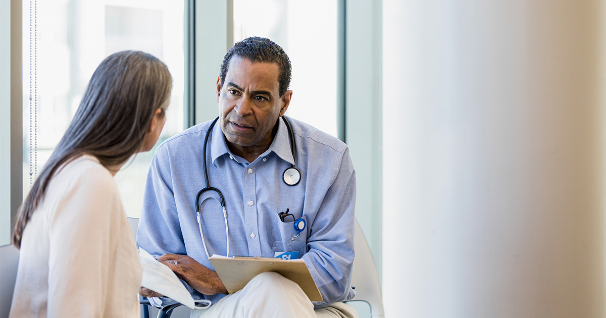 A doctor in a blue shirt wearing a stethoscope listening intently to a female patient in a white blouse.